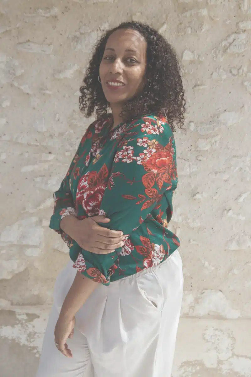 Stecy Aratus, doula de fin de vie dans les Yvelines, souriante, portrait en blouse fleurie devant un mur en pierre